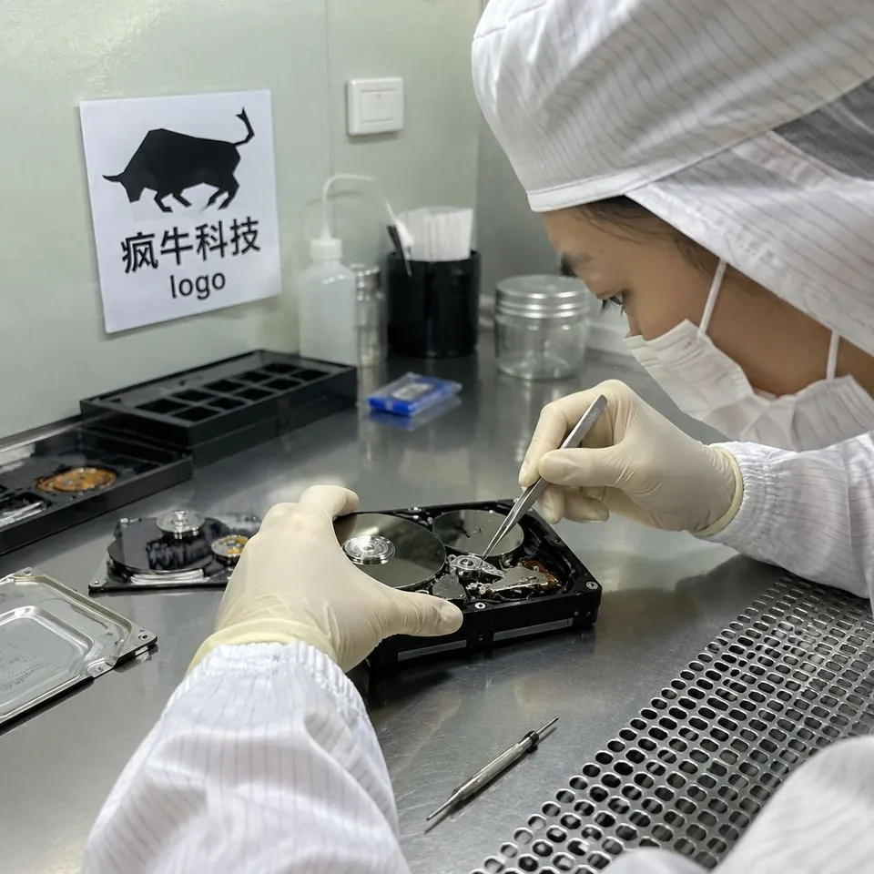 手机纪实抓拍,微距视角。在明亮的万级无尘净化工作台内,一位穿着白色防静电无尘服、戴着乳胶手套的东亚技术员,正使用精密金属镊子小心翼翼地更换一块拆开的3.5寸机械硬盘的磁头。反光的银色盘片上折射出微弱的光芒。工作台侧面贴着一张醒目的「疯牛科技logo」标识贴纸。画面充满真实的硬核维修细节,自然光影,无摆拍感。Authentic candid photography, shot on smartphone, natural daylight, real life repair scene, unposed, slight motion blur, casual perspective, authentic human textures.