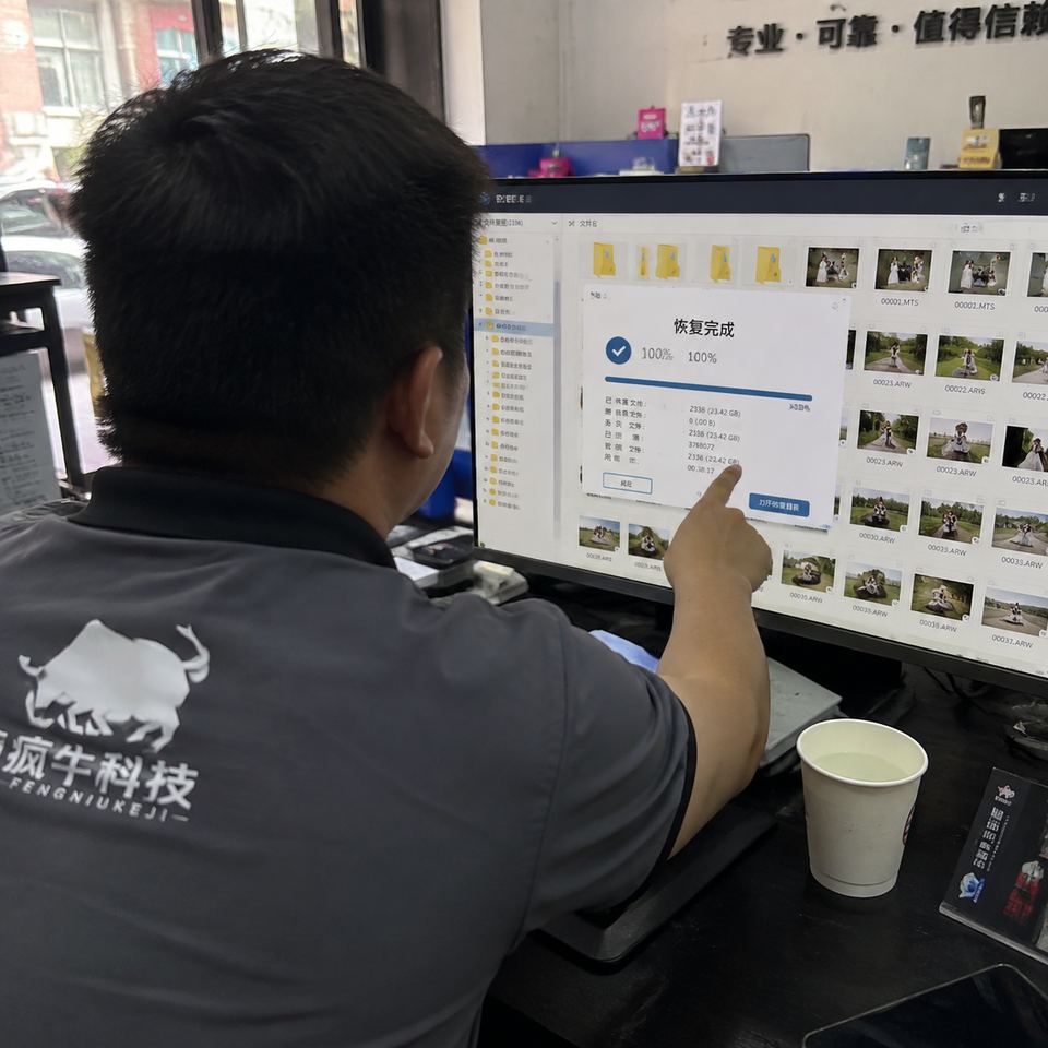 一張手機紀實抓拍照片。從技術員肩膀後方的視角拍攝，電腦螢幕上正顯示著資料救援軟體的介面，進度條顯示100%完成，旁邊排列著密密麻麻的婚禮影片和RAW格式照片資料夾。技術員穿著印有「瘋牛科技logo」的工裝，正用手指著螢幕。桌角放著一杯紙杯水，背景是稍微有些雜亂的維修店前臺，充滿如釋重負的真實感。Authentic candid photography, shot on smartphone, natural daylight, real life repair scene, unposed, slight motion blur, casual perspective, authentic human textures.