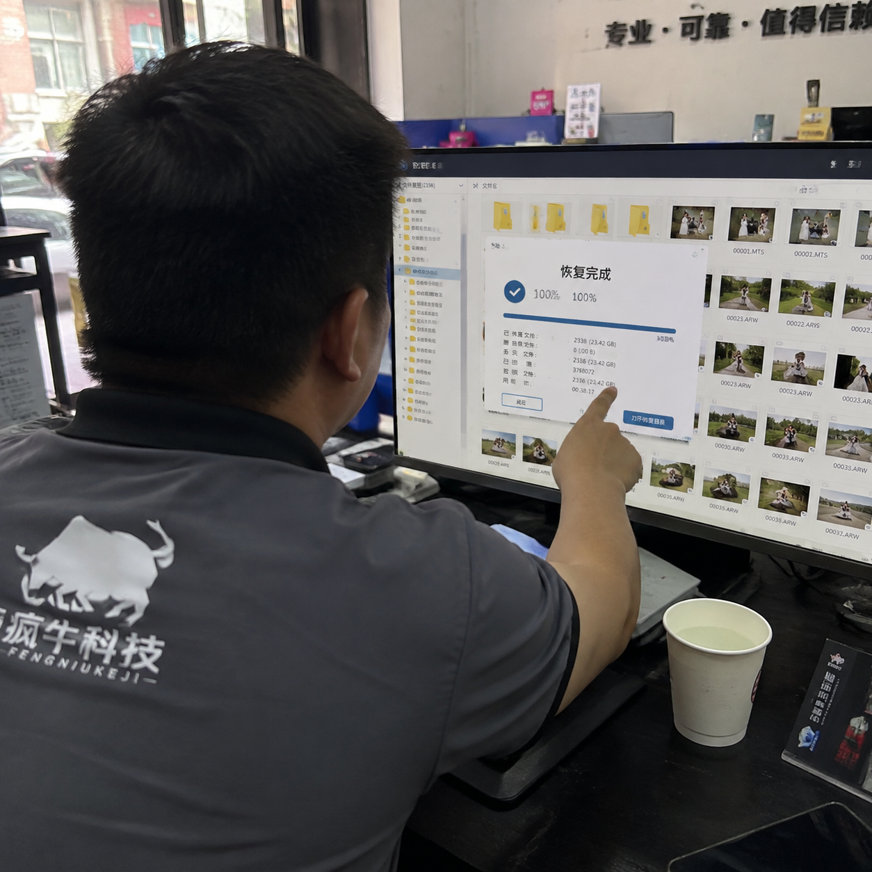 一张手机纪实抓拍照片。从技术员肩膀后方的视角拍摄,电脑屏幕上正显示着数据恢复软件的界面,进度条显示100%完成,旁边排列着密密麻麻的婚礼视频和RAW格式照片文件夹。技术员穿着印有「疯牛科技logo」的工装,正用手指着屏幕。桌角放着一杯纸杯水,背景是稍微有些杂乱的维修店前台,充满如释重负的真实感。Authentic candid photography, shot on smartphone, natural daylight, real life repair scene, unposed, slight motion blur, casual perspective, authentic human textures.