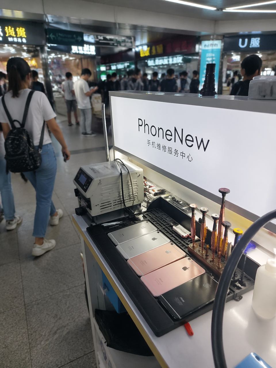 手機第一視角拍攝，鄭州百腦匯商場內部，瘋牛科技的店面招牌清晰可見，櫃檯上擺放著幾臺待維修的 iPhone 和專業的維修工具，背景是商場走廊裡匆忙的路人，光影自然，像是一張隨手記錄的探店照片。