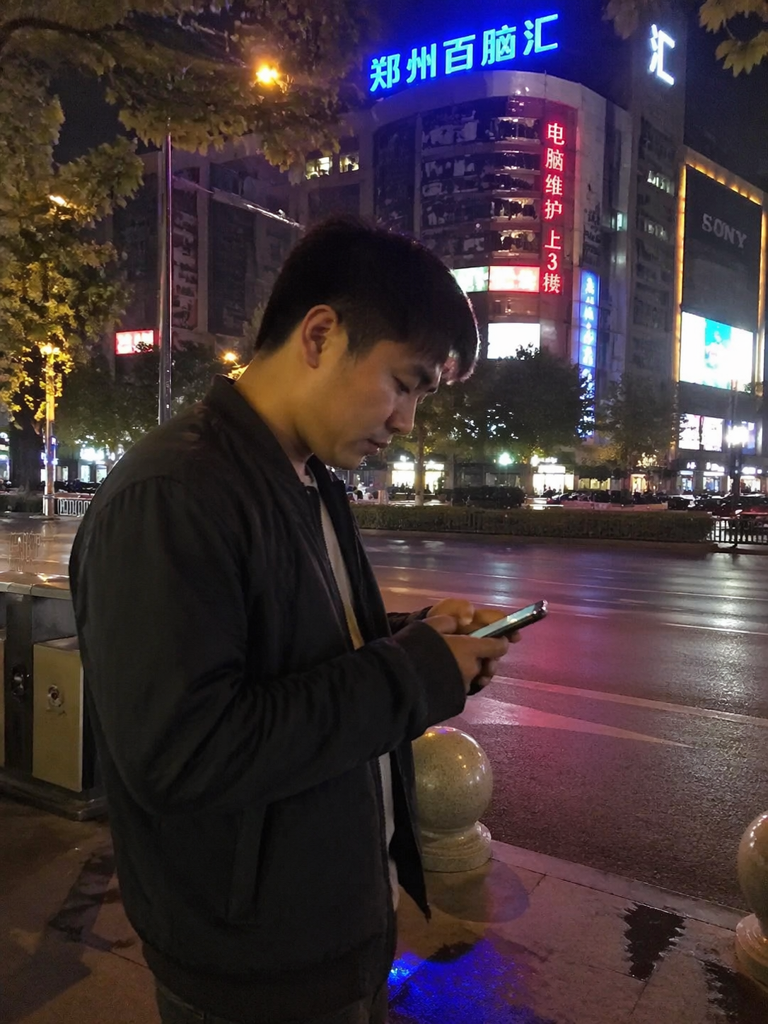 一张自媒体风格的侧拍照片。张铭扬(一位年轻的中国男性创业者)穿着深色休闲夹克,深夜站在郑州百脑汇商场外的街道边,手里拿着手机正在回复消息。背景是深夜街道微弱的霓虹灯光,画面有轻微的手机拍摄虚化感,非常接地气。