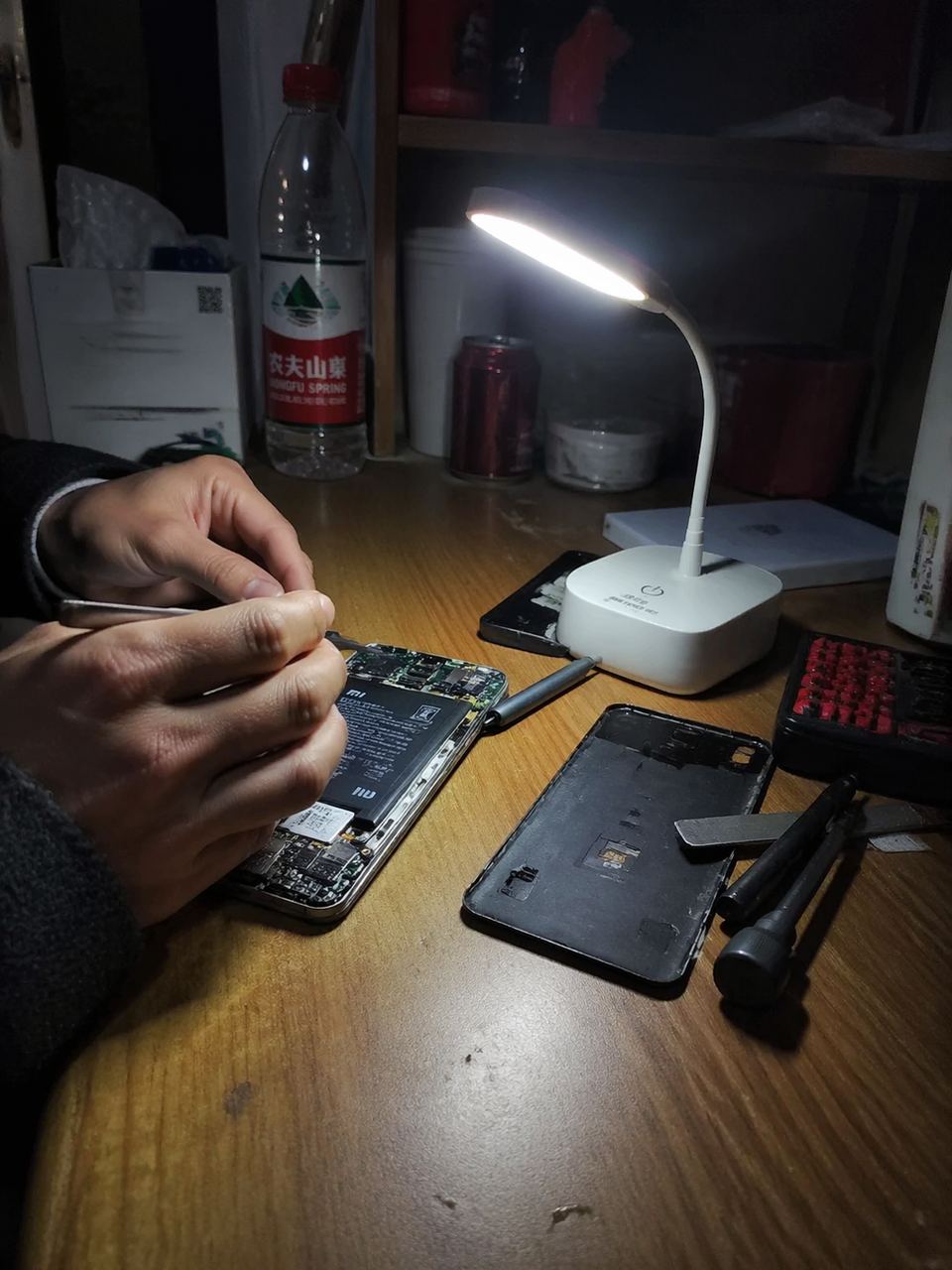 手機隨手拍視角。在昏暗的宿舍內,一張簡陋的木桌上擺放著拆解開的紅米手機,維修師傅的手正在用鑷子小心操作。旁邊放著一個行動式小檯燈,光線集中在主機板上,背景是生活化的雜物,充滿真實的紀實感。
