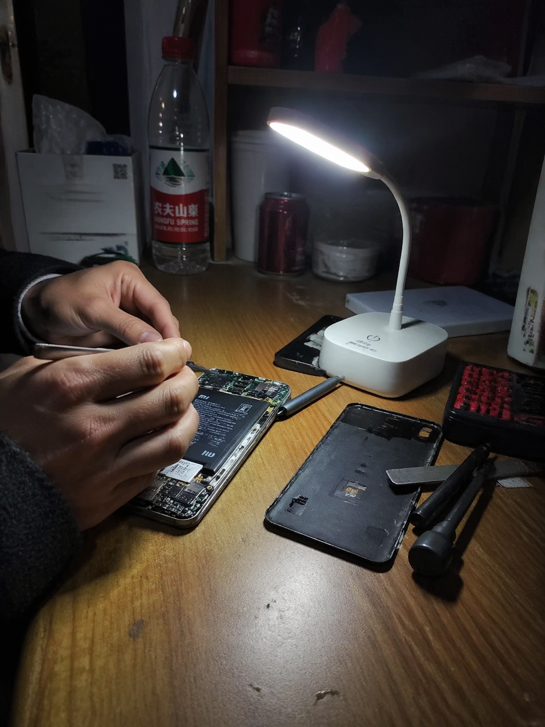 手机随手拍视角。在昏暗的宿舍内,一张简陋的木桌上摆放着拆解开的红米手机,维修师傅的手正在用镊子小心操作。旁边放着一个便携式小台灯,光线集中在主板上,背景是生活化的杂物,充满真实的纪实感。