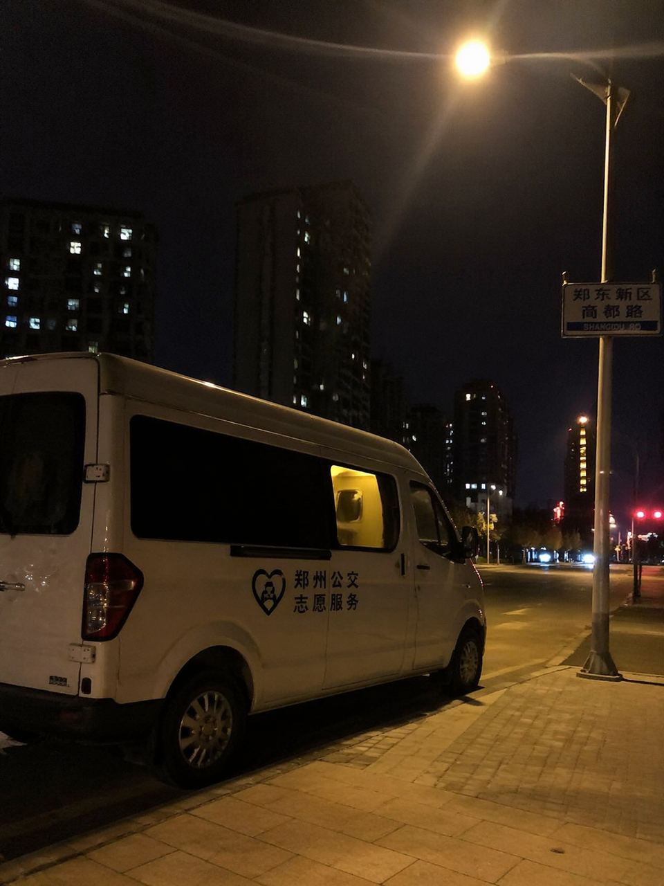 溫暖的紀實畫面。深夜兩點的鄭州街頭,一輛貼有簡約標識的白色服務車停在路燈下,車內透出微弱的暖色燈光。畫面構圖隨意自然,帶有生活化的親和力,展現出深夜守護的真實氛圍。