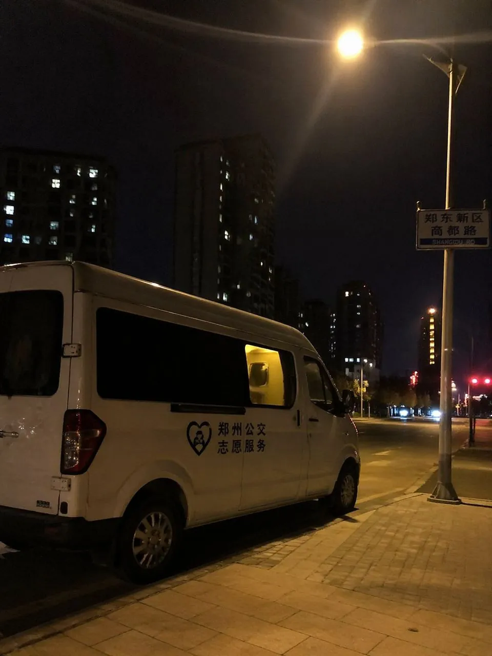 温暖的纪实画面。深夜两点的郑州街头,一辆贴有简约标识的白色服务车停在路灯下,车内透出微弱的暖色灯光。画面构图随意自然,带有生活化的亲和力,展现出深夜守护的真实氛围。