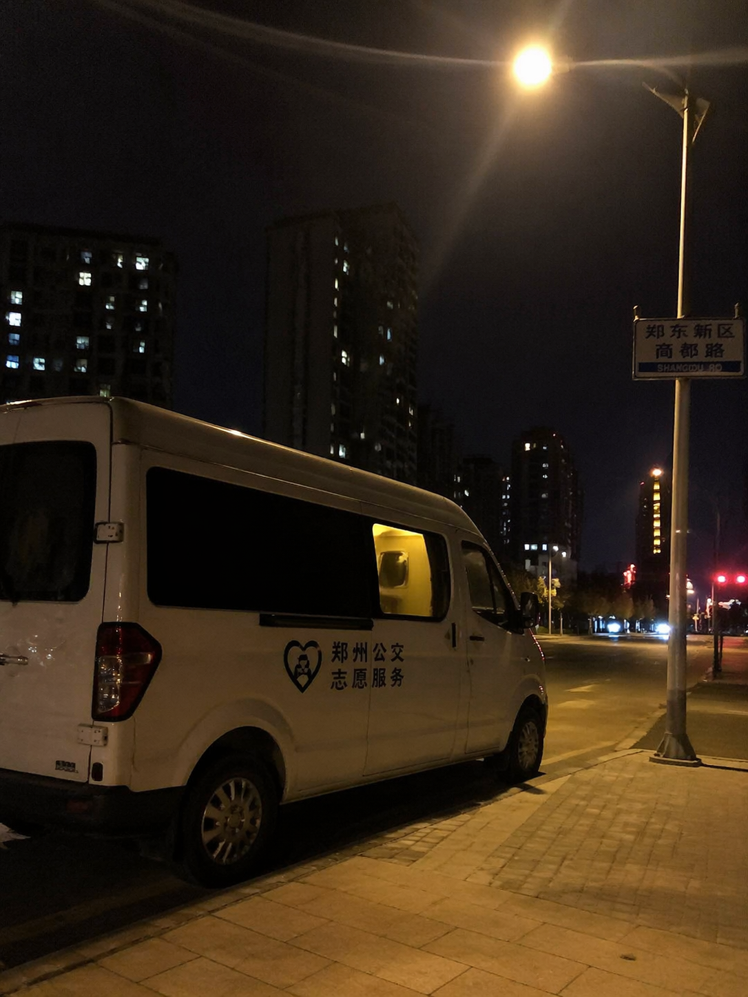温暖的纪实画面。深夜两点的郑州街头,一辆贴有简约标识的白色服务车停在路灯下,车内透出微弱的暖色灯光。画面构图随意自然,带有生活化的亲和力,展现出深夜守护的真实氛围。