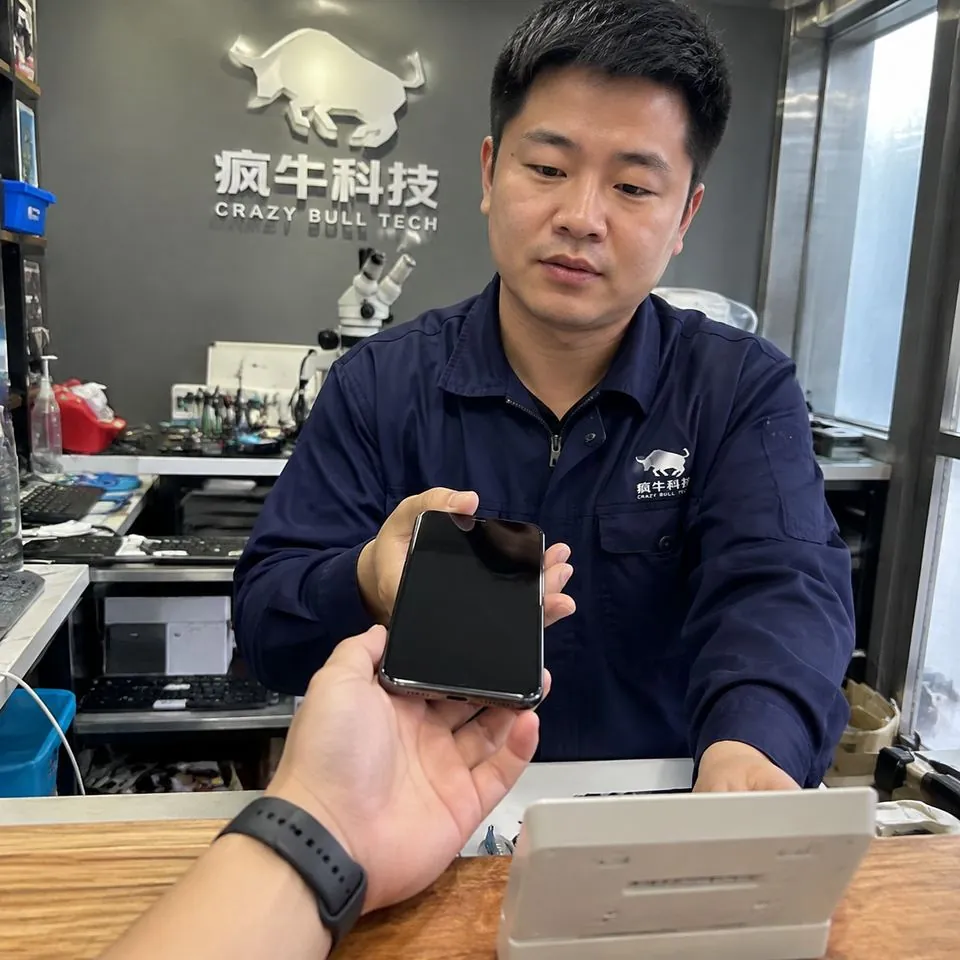 手机纪实抓拍镜头。一位顾客焦虑的手正将一部黑屏的iPhone递过木质维修柜台，柜台对面是一位穿着深蓝色工作服的东亚男技术员。背景墙上隐约可见“疯牛科技logo”的品牌标识。自然光线从侧面窗户照入，画面带有一点生活化的杂乱感，真实的皮肤纹理，无摆拍感。Authentic candid photography, shot on smartphone, natural daylight, real life repair scene, unposed, slight motion blur, casual perspective, authentic human textures.