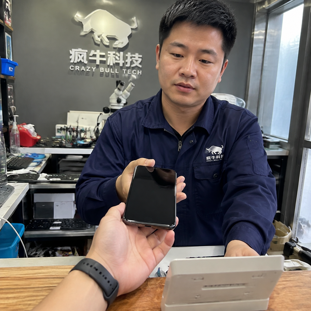 手机纪实抓拍镜头。一位顾客焦虑的手正将一部黑屏的iPhone递过木质维修柜台,柜台对面是一位穿着深蓝色工作服的东亚男技术员。背景墙上隐约可见“疯牛科技logo”的品牌标识。自然光线从侧面窗户照入,画面带有一点生活化的杂乱感,真实的皮肤纹理,无摆拍感。Authentic candid photography, shot on smartphone, natural daylight, real life repair scene, unposed, slight motion blur, casual perspective, authentic human textures.