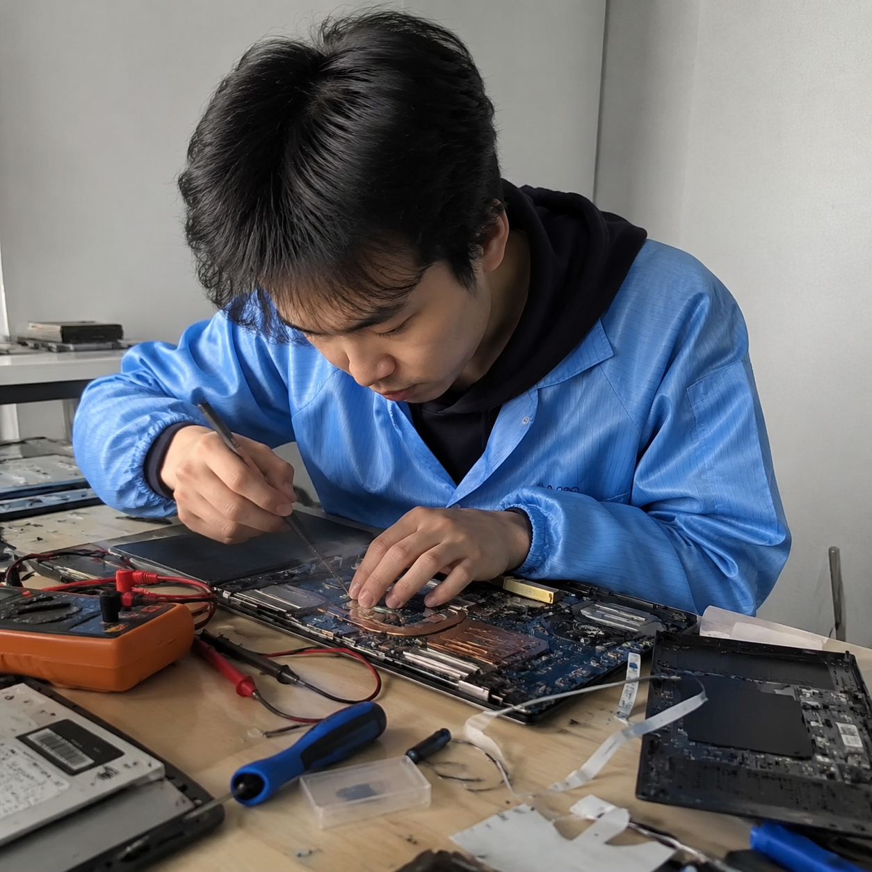 手机纪实抓拍镜头,一位东亚年轻男技术员穿着蓝色防静电工装,在稍微有些杂乱但专业的维修工作台前专注工作。桌上有一台拆开的笔记本电脑,露出精密的主板芯片,旁边散落着万用表、螺丝刀和排线。自然光从侧面照亮工作台,画面具有真实的生活气息和轻微的抓拍动感。Authentic candid photography, shot on smartphone, natural daylight, real life repair scene, unposed, slight motion blur, casual perspective, authentic human textures.