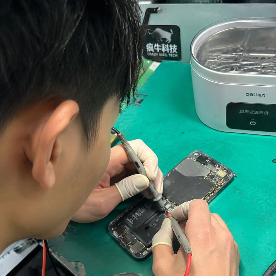 手机随手拍的微距特写镜头。维修台的绿色静电垫上,一只戴着指套的东亚技术人员的手,正拿着精细的金属探针和万用表表笔,检测一部拆开的iPhone手机充电接口触点。旁边放着一台小型超声波清洗仪,背景里隐约可见工作台一角贴着的「疯牛科技logo」的标识贴纸。自然光线,焦点在主板的微小芯片和铜绿腐蚀点上,画面真实自然,带有一点生活化的杂乱感。Authentic candid photography, shot on smartphone, natural daylight, real life repair scene, unposed, slight motion blur, casual perspective, authentic human textures.