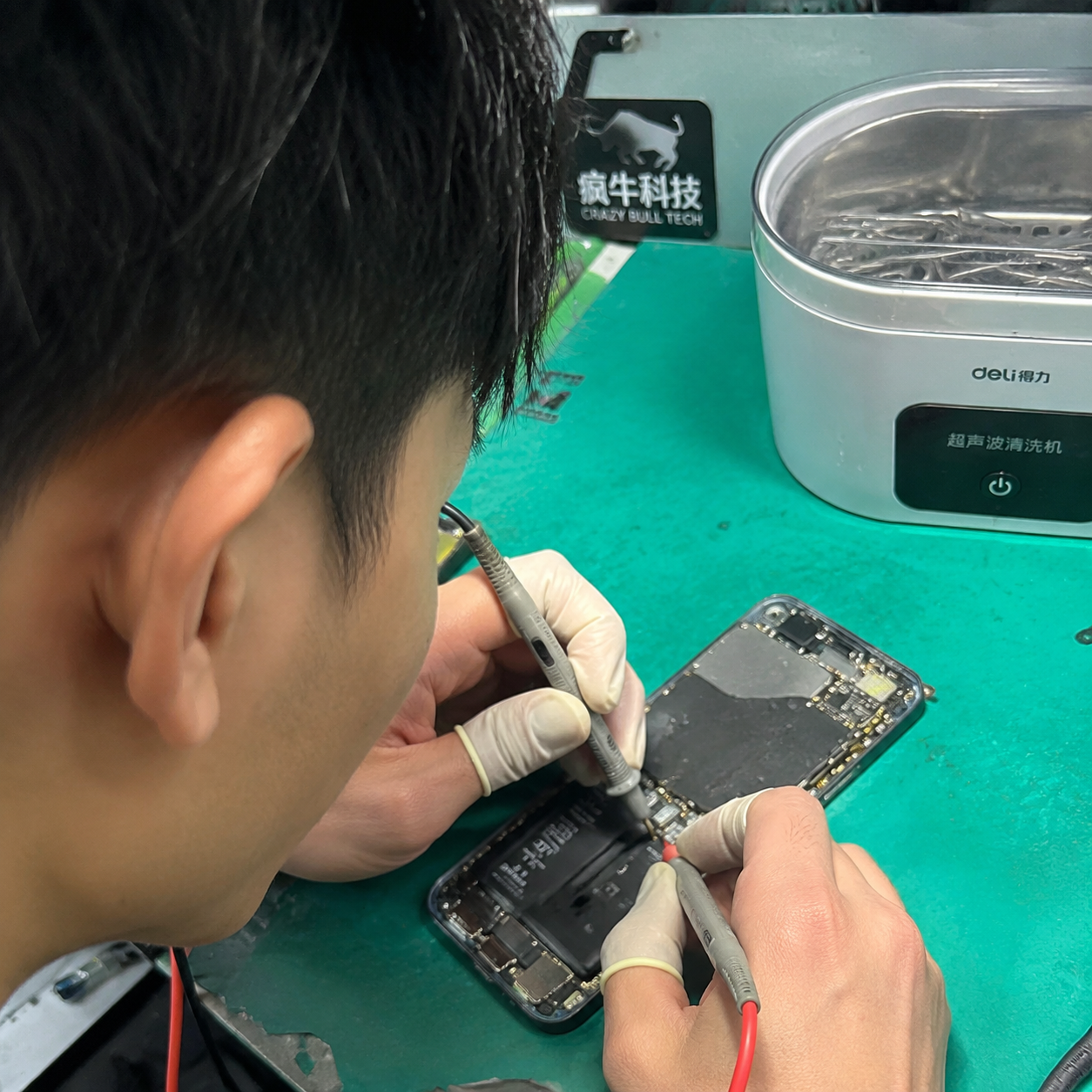 手机随手拍的微距特写镜头。维修台的绿色静电垫上,一只戴着指套的东亚技术人员的手,正拿着精细的金属探针和万用表表笔,检测一部拆开的iPhone手机充电接口触点。旁边放着一台小型超声波清洗仪,背景里隐约可见工作台一角贴着的「疯牛科技logo」的标识贴纸。自然光线,焦点在主板的微小芯片和铜绿腐蚀点上,画面真实自然,带有一点生活化的杂乱感。Authentic candid photography, shot on smartphone, natural daylight, real life repair scene, unposed, slight motion blur, casual perspective, authentic human textures.
