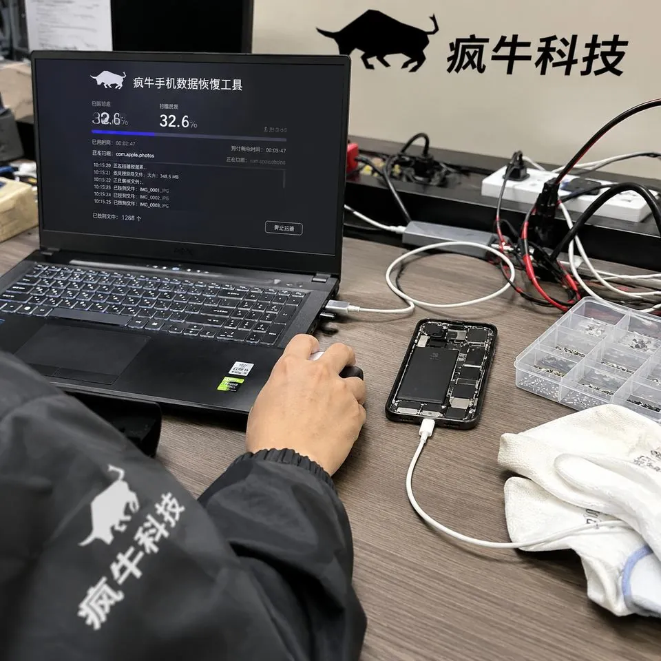 手机纪实抓拍感。凌乱而专业的电子数据恢复工作台实拍。一台笔记本电脑屏幕上正运行着“疯牛手机数据恢复工具”的扫描界面,旁边用数据线连接着一部拆开后盖的智能手机。一位穿着“疯牛科技”Logo防静电服的东亚技术员的一只手正在操控鼠标。工作台上散落着测试线缆、防静电手套和螺丝盒。自然光,iPhone随手拍视角,充满生活气息与极强的真实信任感。