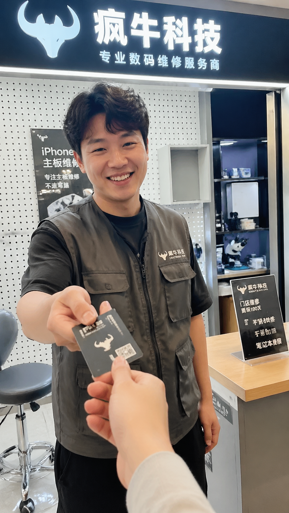 生活化纪实,写实照片。在一家位于中国商场内的现代数码维修门店前,一位身穿印有「疯牛科技logo」工装的店员正微笑着递给顾客一张名片,背景可以看到整洁的维修柜台和「疯牛科技」的品牌灯箱。光影柔和自然,具有极强的亲和力和真实感。Authentic candid photography, shot on smartphone, social media vlog style, natural daylight, unposed, real life scene, slight motion blur, casual perspective, 35mm镜头拍摄。